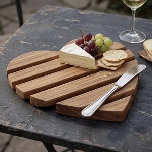 May include: A heart-shaped wooden serving board made of horizontal slats, featuring a wedge of brie cheese, a cluster of red and green grapes, three round crackers, and a silver-toned metal spreader knife resting on the surface.
