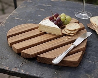 May include: A heart-shaped wooden serving board made of horizontal slats, featuring a wedge of brie cheese, a cluster of red and green grapes, three round crackers, and a silver-toned metal spreader knife resting on the surface.