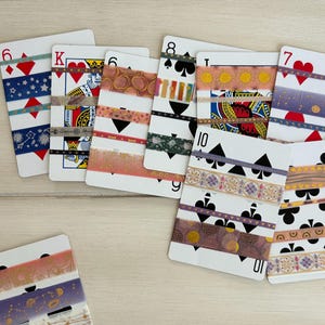 May include: A collection of decorative washi tape strips applied to standard playing cards. The tapes feature various patterns including celestial motifs, geometric shapes, floral designs, and metallic accents in shades of gold, pink, blue, and black.