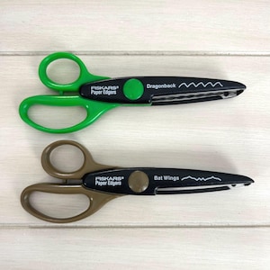 May include: Two pairs of Fiskars Paper Edgers scissors with black blades and colored handles. The top pair has bright green handles and is labeled Dragonback with a wavy line icon. The bottom pair has tan handles and is labeled Bat Wings with a jagged line icon.