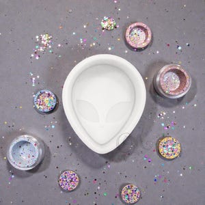 May include: A white silicone alien head mold surrounded by small, round containers filled with colorful glitter. The glitter is in various colors, including pink, purple, and blue, scattered on a gray surface. The mold has a smooth finish.
