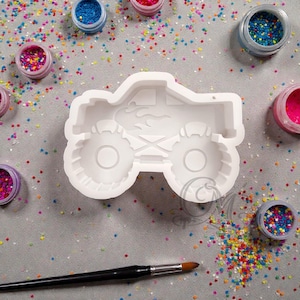 May include: A white silicone mold in the shape of a monster truck with large tires and flame details. The mold is surrounded by small jars of colorful glitter and a paintbrush, suggesting a crafting or DIY theme.
