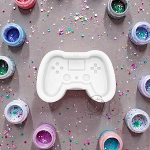 May include: A white silicone mold in the shape of a video game controller is surrounded by small jars filled with colorful glitter. The glitter is in shades of pink, blue, green, and purple. The background is a neutral gray.