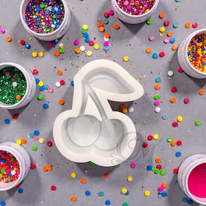 May include: A white silicone cherry-shaped mold with a stem and two cherries. The mold is surrounded by small containers filled with colorful glitter and confetti, scattered on a gray surface.