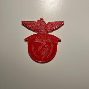 Może przedstawiać: Czerwony emblemat przedstawiający stylizowanego orła z rozpostartymi skrzydłami nad okrągłym wzorem. Emblemat ma teksturowaną powierzchnię i wydaje się być wykonany z litego materiału. Projekt jest symbolem.