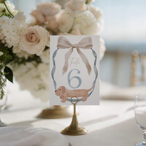 May include: A table number card with a watercolor illustration of a dachshund dog and a bow. The card reads "Table 6" in elegant script. The card is held in a gold-colored metal stand, with a floral arrangement in the background.