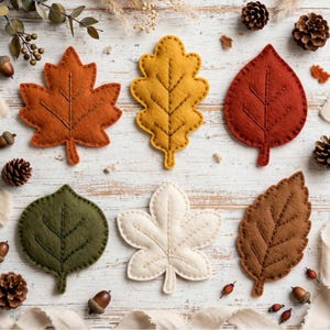 Può includere: Un set di sei foglie in feltro di varie forme e colori, tra cui arancione, giallo, rosso, verde oliva, crema e marrone. Ogni foglia presenta dettagli di venature cucite e una consistenza morbida e imbottita su una superficie in legno bianco.