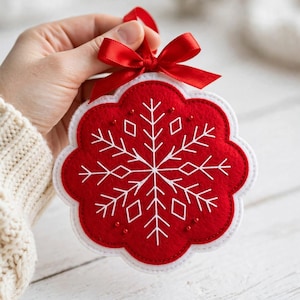 Puede incluir: Un adorno de fieltro rojo con forma circular festoneada, que presenta detalles de copos de nieve bordados en blanco y pequeños abalorios rojos. El adorno está rematado con un lazo de satén rojo para colgarlo.
