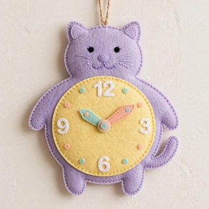May include: A lavender felt wall clock shaped like a cat with a yellow circular face. The clock features white embroidered numbers 3, 6, 9, and 12, small teal and peach dots for markers, and teal and peach clock hands. A string is attached to the top.