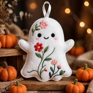 Puede incluir: Un adorno de fantasma de fieltro blanco con una cinta para colgar, que presenta detalles bordados que incluyen dos ojos negros, una pequeña sonrisa, mejillas rosadas y tallos florales coloridos con flores rojas y rosas y hojas verdes.