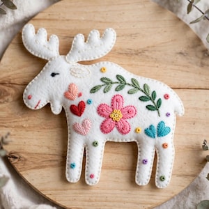 May include: A white felt moose ornament featuring hand-stitched embroidery. The design includes a large pink flower, green leafy branches, and several small hearts in shades of red, pink, and blue, accented with colorful French knot details.