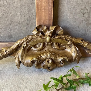 Könnte beinhalten: Ein goldfarbenes dekoratives Wandornament mit einem zentralen Schleifenmotiv, fließenden Bändern und kunstvoll geschnitzten Blattgirlanden, die sich zu beiden Seiten erstrecken. Das Stück hat eine verwitterte Metalloberfläche.