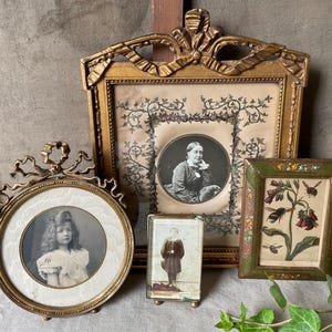 May include: A collection of antique picture frames in various shapes and sizes. The frames are gold-toned and ornate, holding vintage photographs and botanical prints. The frames are arranged on a neutral-toned surface.