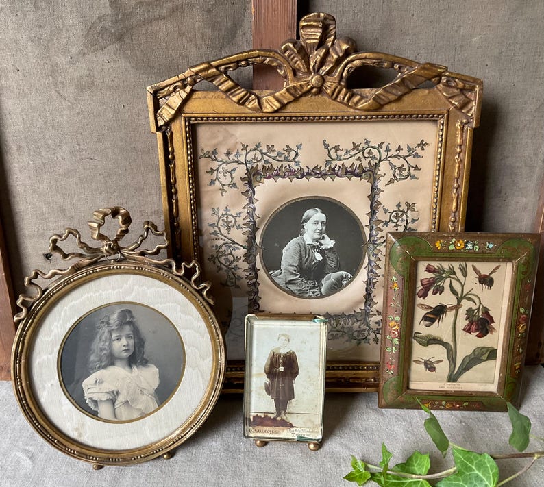 May include: A collection of antique picture frames in various shapes and sizes. The frames are gold-toned and ornate, holding vintage photographs and botanical prints. The frames are arranged on a neutral-toned surface.