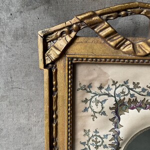 May include: Antique gold-colored picture frame with ornate detailing. The frame surrounds a portrait with floral embellishments in shades of blue, green, and purple. The frame's design includes ribbon-like accents.