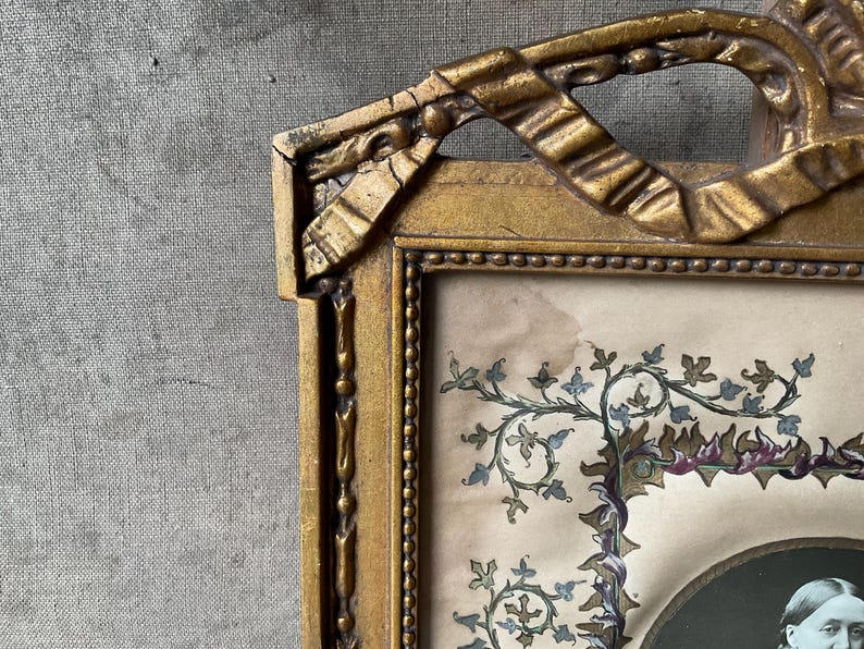 May include: Antique gold-colored picture frame with ornate detailing. The frame surrounds a portrait with floral embellishments in shades of blue, green, and purple. The frame's design includes ribbon-like accents.