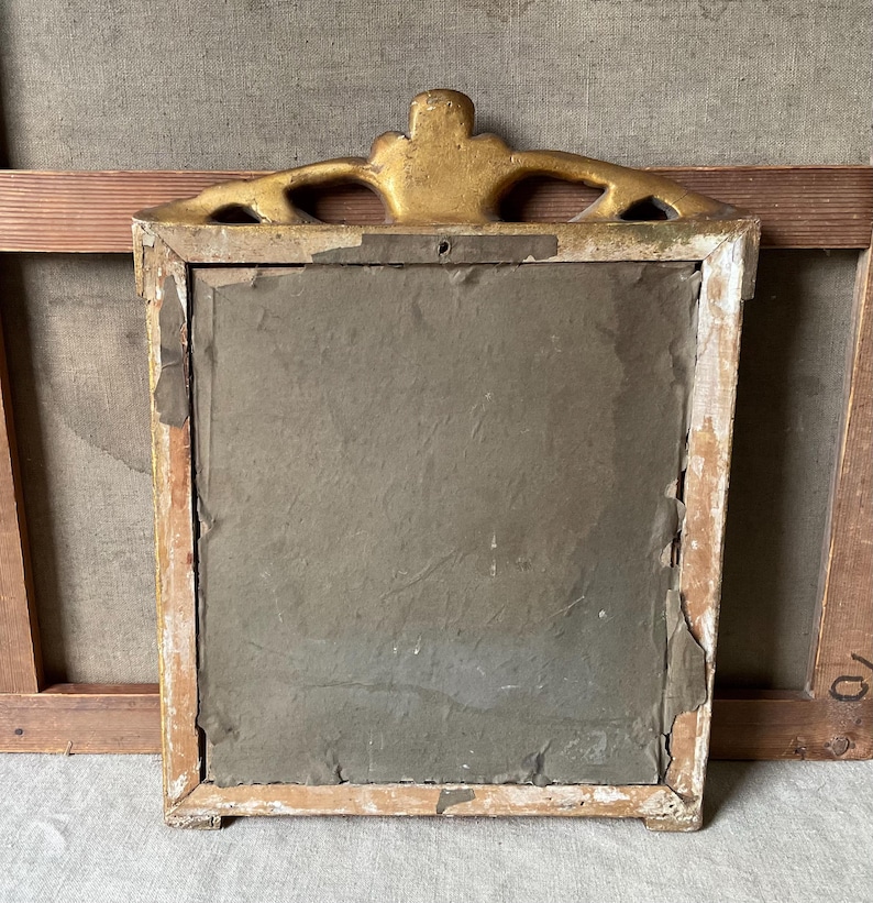 May include: Back of a vintage rectangular framed artwork. The frame is painted white with gold accents at the top. The artwork is covered with a gray paper backing. The frame is set against a neutral background.