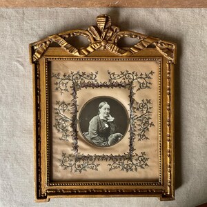 May include: An antique gold-colored picture frame with a portrait of a woman. The frame has ornate detailing and a decorative bow at the top. The portrait is in a circular frame with floral embellishments.