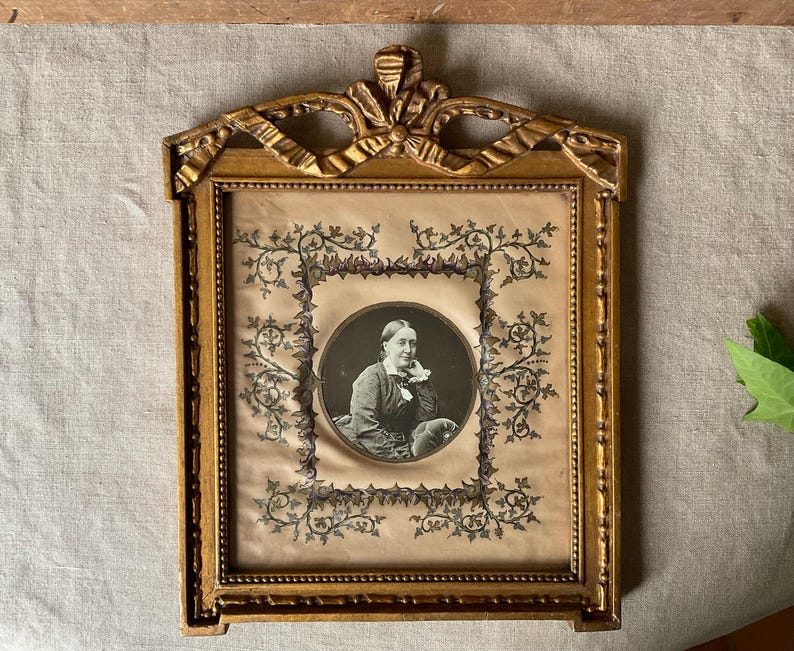 May include: An antique gold-colored picture frame with a portrait of a woman. The frame has ornate detailing and a decorative bow at the top. The portrait is in a circular frame with floral embellishments.