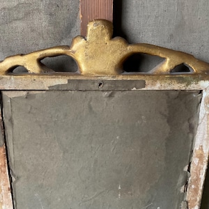 May include: An antique, rectangular mirror frame with a gold-painted, ornate top. The frame is made of wood, with visible signs of age and wear, including chipped paint and a distressed finish. The mirror's surface is a muted gray.
