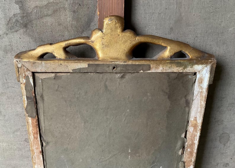 May include: An antique, rectangular mirror frame with a gold-painted, ornate top. The frame is made of wood, with visible signs of age and wear, including chipped paint and a distressed finish. The mirror's surface is a muted gray.