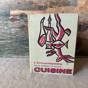 Op de afbeelding: Een hardcover boek getiteld L'Encyclopédie de la Grande et Petite Cuisine met een getextureerde gebroken witte kaft. De voorzijde toont een gestileerde grafische illustratie in zwart en magenta van een fles, een karaf en een vis.
