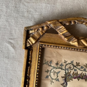May include: A vintage gold-colored picture frame with ornate detailing. The frame has a ribbon-like design at the top and a beaded border. The artwork inside features a botanical illustration with green, blue, and maroon accents.