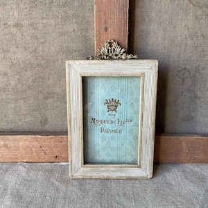 Può includere: Cornice rettangolare in stile antico con bordo chiaro e strutturato. La cornice contiene una stampa blu-verde con il testo "Marque de Fabrique Déposée Paris" e un'emblema a corona. Un accento decorativo color oro si trova nella parte superiore.