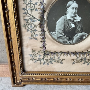 May include: Antique portrait of a woman in a gold ornate frame. The black and white portrait is oval-shaped, surrounded by a decorative border with floral and vine motifs in blue, green, and purple. The frame is gold.