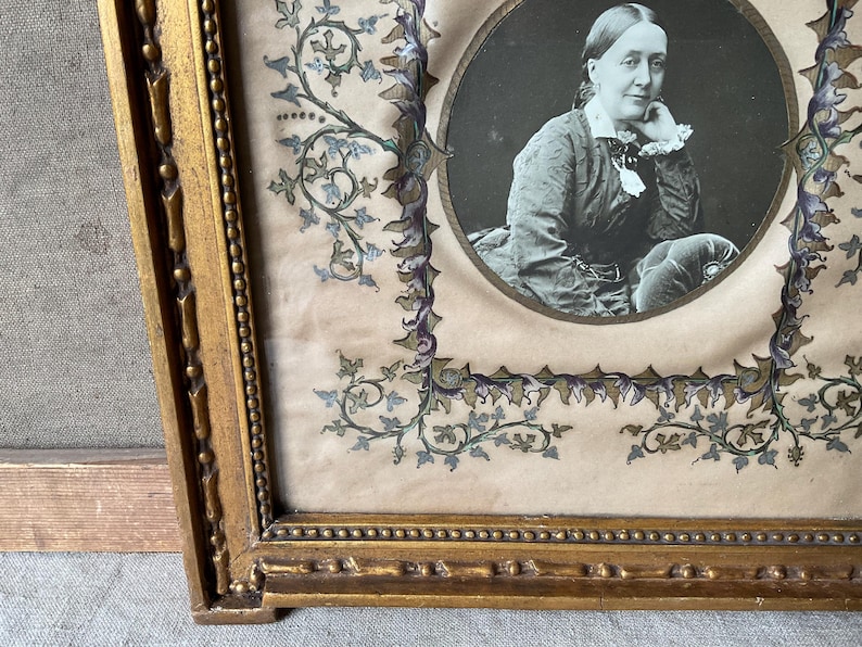 May include: Antique portrait of a woman in a gold ornate frame. The black and white portrait is oval-shaped, surrounded by a decorative border with floral and vine motifs in blue, green, and purple. The frame is gold.