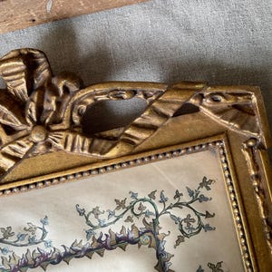 May include: Ornate gold-colored picture frame with a decorative bow at the top. The frame surrounds a print with a floral and vine design in shades of green, purple, and blue. The frame has a beaded border.