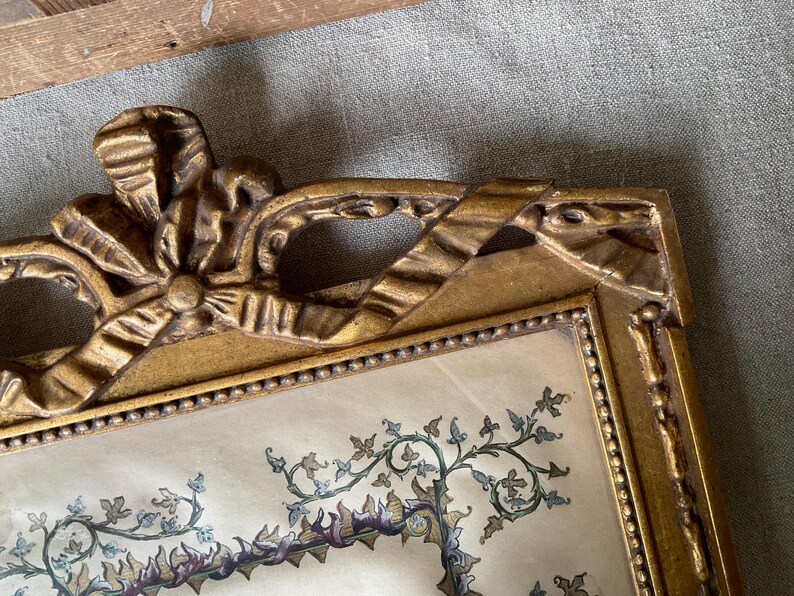 May include: Ornate gold-colored picture frame with a decorative bow at the top. The frame surrounds a print with a floral and vine design in shades of green, purple, and blue. The frame has a beaded border.