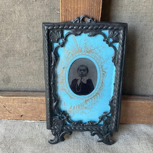 May include: An antique black metal picture frame with a portrait of a person. The frame has ornate detailing and a light blue background with gold accents. The portrait is oval-shaped and in black and white.