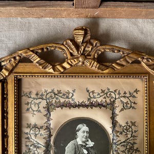 May include: An antique portrait in a gold-colored ornate frame. The portrait features a woman in a dark dress, set against a light background. The frame has a decorative top with ribbon and bow details, and the portrait is surrounded by floral embellishments.