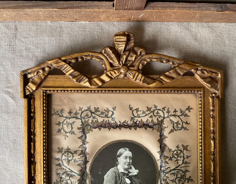 May include: An antique portrait in a gold-colored ornate frame. The portrait features a woman in a dark dress, set against a light background. The frame has a decorative top with ribbon and bow details, and the portrait is surrounded by floral embellishments.
