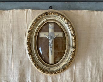 May include: An oval wall-mounted frame with a cream-colored finish and gold-toned ornate detailing. Inside the glass-covered frame, a light-colored cross features a silver-toned metal crucifix figure with a sunburst halo and a small silver plaque.