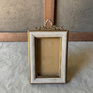 May include: A rectangular picture frame with a gold-colored border and a white and gold striped inner frame. The top of the frame features a decorative gold-colored wreath. The frame is empty and ready for a photo.