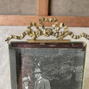 Pode incluir: Uma moldura retangular de vidro com um topo decorativo de latão em forma de laço. A moldura contém uma fotografia a preto e branco de duas pessoas. O ornamento de latão está fixado na borda superior do vidro espesso da moldura.