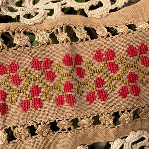 Puede incluir: Primer plano de una tela beige con bordado de punto de cruz floral rojo y tallos verdes. La tela está bordeada con un ribete calado decorativo y un encaje color crema.