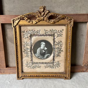 May include: Antique gold-colored framed portrait. The frame has a decorative bow at the top and intricate detailing. The portrait is a black and white image of a person in a circular frame, surrounded by floral designs.