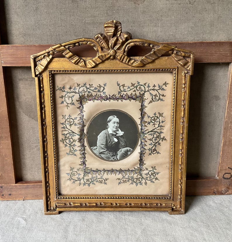 May include: Antique gold-colored framed portrait. The frame has a decorative bow at the top and intricate detailing. The portrait is a black and white image of a person in a circular frame, surrounded by floral designs.