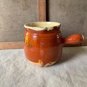 Może przedstawiać: Okrągły ceramiczny garnek z błyszczącym, głębokim czerwonobrązowym szkliwem na zewnątrz i kremowym wnętrzem. Naczynie posiada pojedynczy wystający uchwyt z boku, szeroki okrągły otwór oraz grubą, zaokrągloną krawędź z małym dzióbkiem do nalewania.