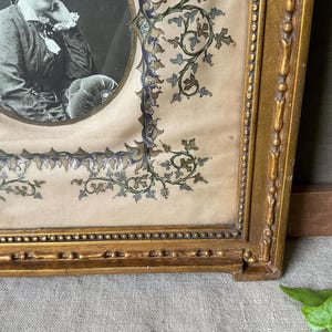 May include: Antique framed portrait featuring a black and white oval photograph. The frame is ornate, gold-colored, and rectangular, with decorative floral and vine accents in blue and green. The portrait is of a person.