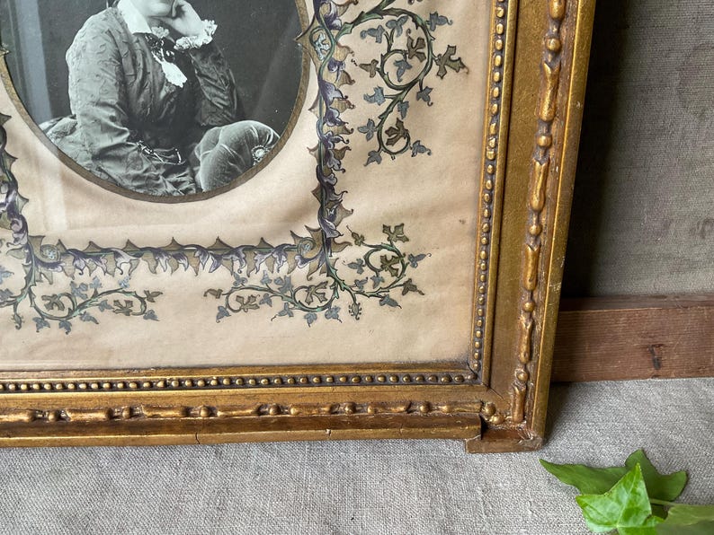May include: Antique framed portrait featuring a black and white oval photograph. The frame is ornate, gold-colored, and rectangular, with decorative floral and vine accents in blue and green. The portrait is of a person.