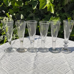 Puede incluir: Seis copas de champán transparentes con tallos, dispuestas sobre un mantel blanco estampado. Las copas tienen diferentes diseños y se colocan sobre un fondo de follaje verde.