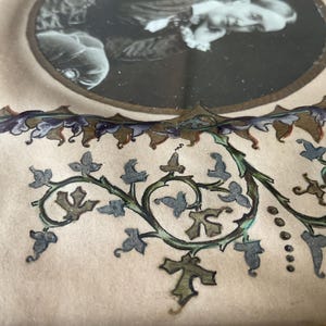 May include: Close-up of a vintage photograph in a decorative frame. The frame features a cream-colored background with a detailed floral design in green, blue, and brown. The photograph is in a circular frame with a gold border.