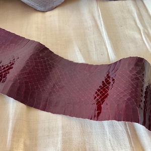 May include: A strip of burgundy-colored snakeskin leather with a glossy finish. The leather has a scale pattern and is curved, with a lighter area on the right side. The material is likely used for crafting.