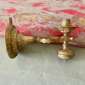 May include: An antique gold-colored wall sconce with ornate detailing. The sconce features a circular backplate, a curved arm, and a candle cup. The design includes floral and scroll patterns, adding a vintage aesthetic. The sconce is made of metal.
