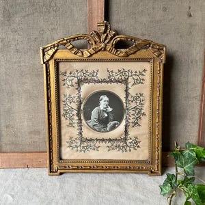 May include: Ornate gold-colored picture frame with a portrait of a person in a black and white oval. The frame has a decorative bow at the top and floral embellishments. The portrait is surrounded by a floral design.