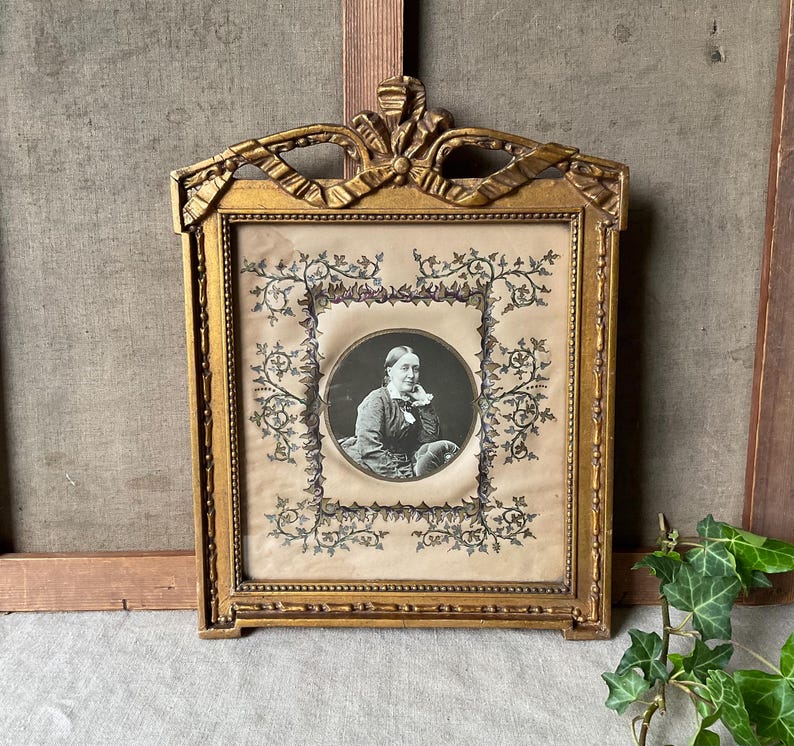 May include: Ornate gold-colored picture frame with a portrait of a person in a black and white oval. The frame has a decorative bow at the top and floral embellishments. The portrait is surrounded by a floral design.
